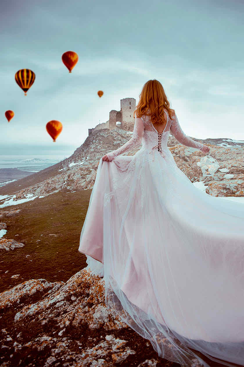 Woman in a blush wedding gown facing castle ruins on a cliff, hot air balloons in the sky