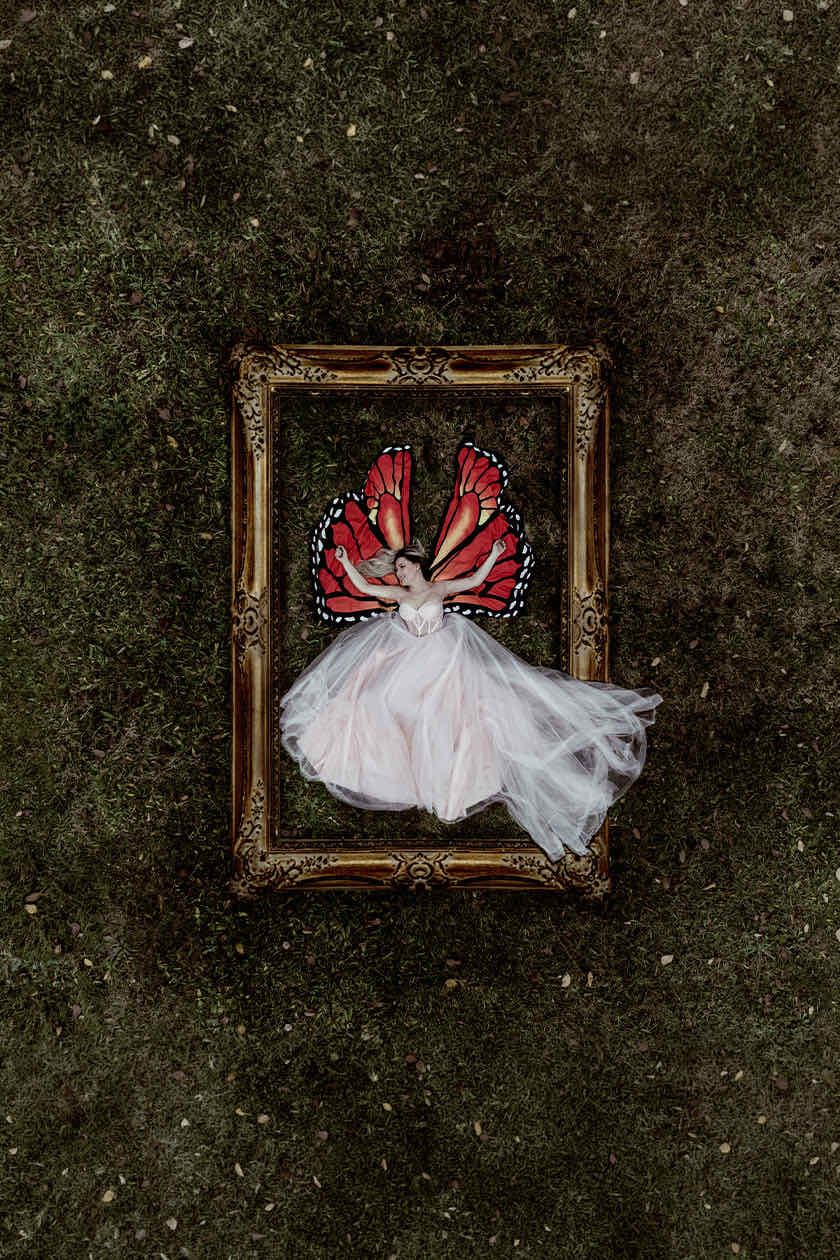 Overhead shot of a woman with butterfly wings lying inside a vintage gold picture frame on grass