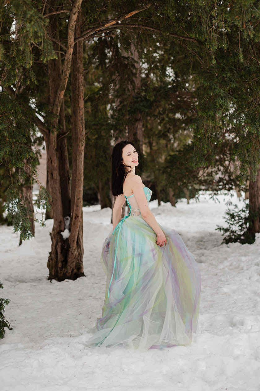 Woman in a pastel multicolor tulle gown smiling over her shoulder in a snowy pine forest